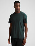 A man wearing a short sleeve green t-shirt and black pants against a plain studio background.