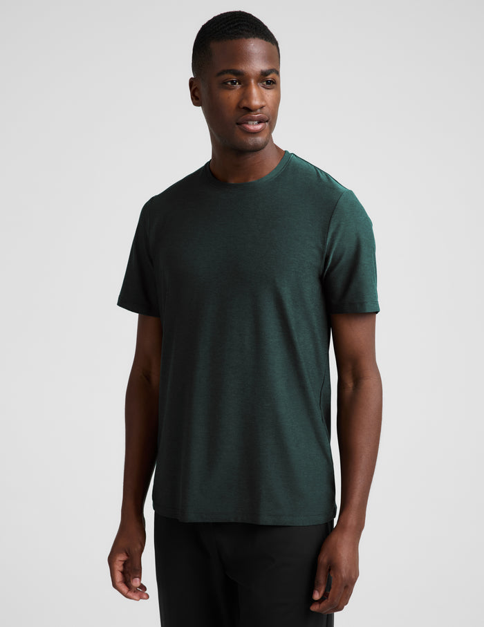 A man wearing a short sleeve green t-shirt and black pants against a plain studio background.