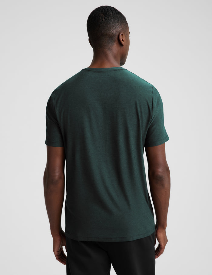 A man wearing a short sleeve green t-shirt and black pants against a plain studio background.