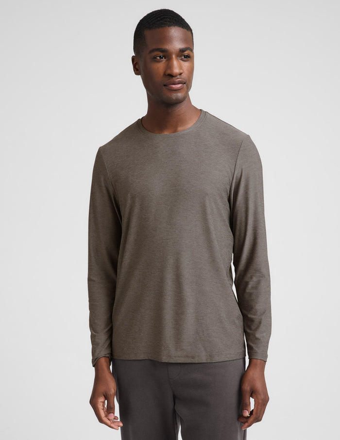 A man wearing a brown long sleeve crew neck shirt and dark gray pants against a plain studio background.