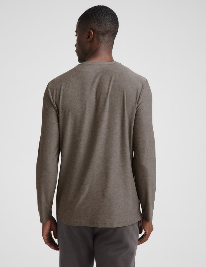 A man wearing a brown long sleeve crew neck shirt and dark gray pants against a plain studio background.