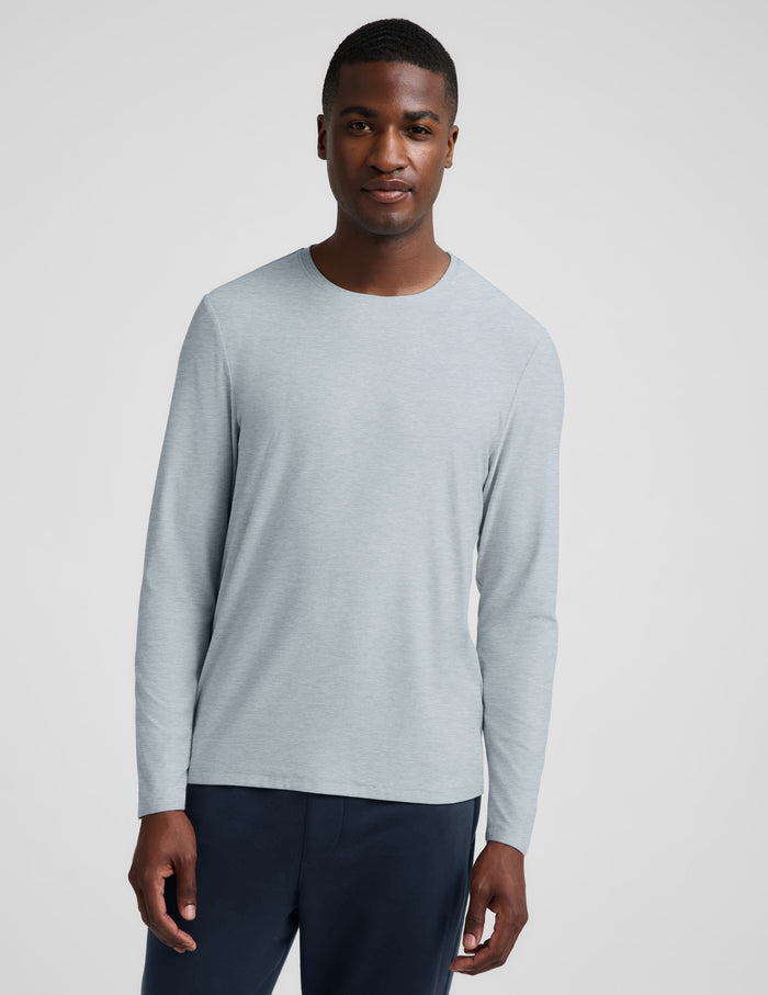 A man wearing a light gray long-sleeve crewneck shirt and dark navy pants, standing against a light gray studio background.