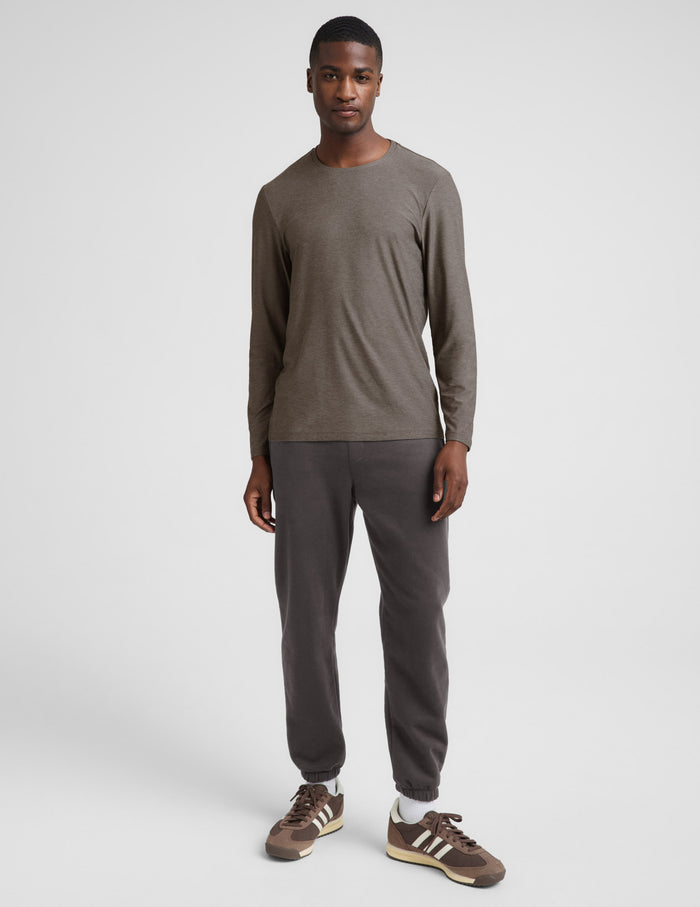 A man wearing a brown long sleeve crew neck shirt and dark gray pants against a plain studio background.