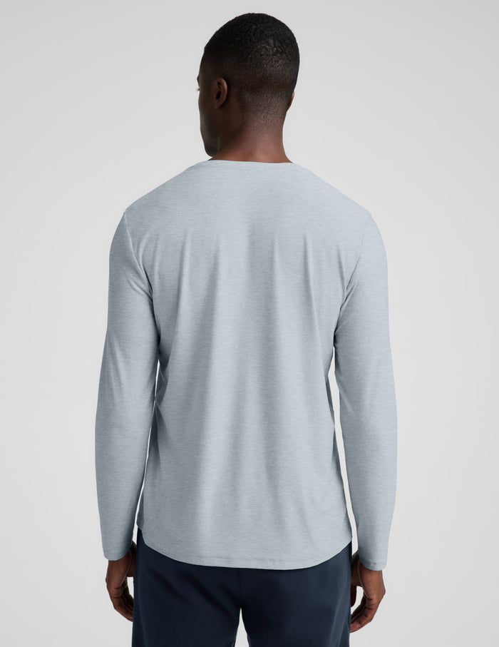 A man wearing a light gray long-sleeve crewneck shirt and dark navy pants, standing against a light gray studio background.