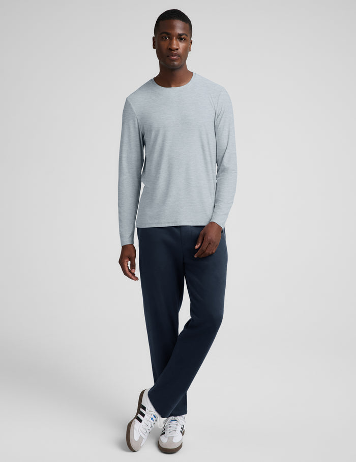 A man wearing a light gray long-sleeve crewneck shirt and dark navy pants, standing against a light gray studio background.