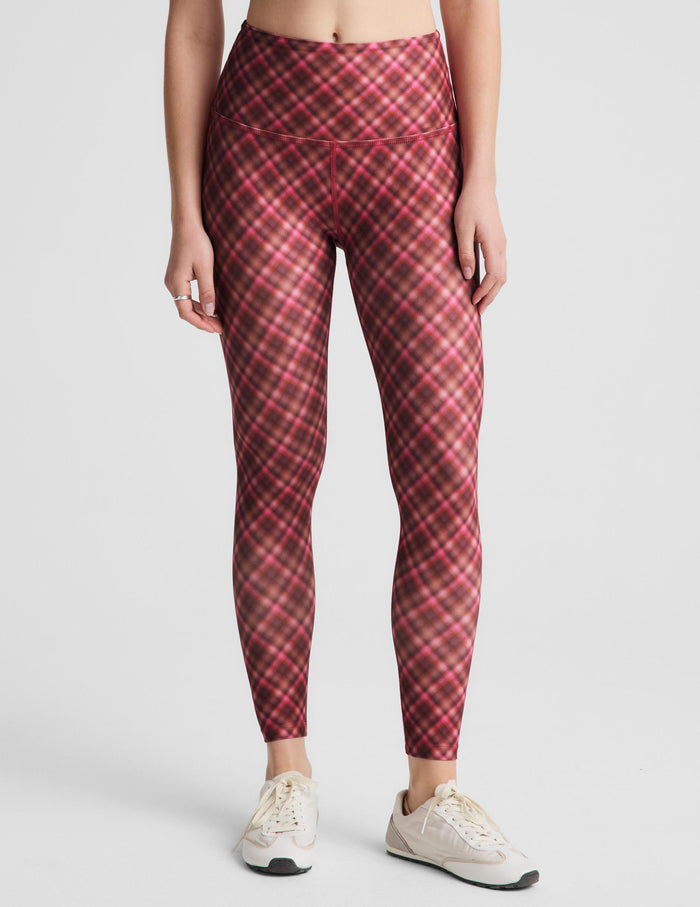 A woman modeling a matching red and pink plaid activewear set, consisting of a sports bra and high-waisted leggings, paired with white sneakers, against a light gray studio background.