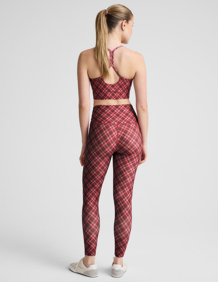 A woman modeling a matching red and pink plaid activewear set, consisting of a sports bra and high-waisted leggings, paired with white sneakers, against a light gray studio background.