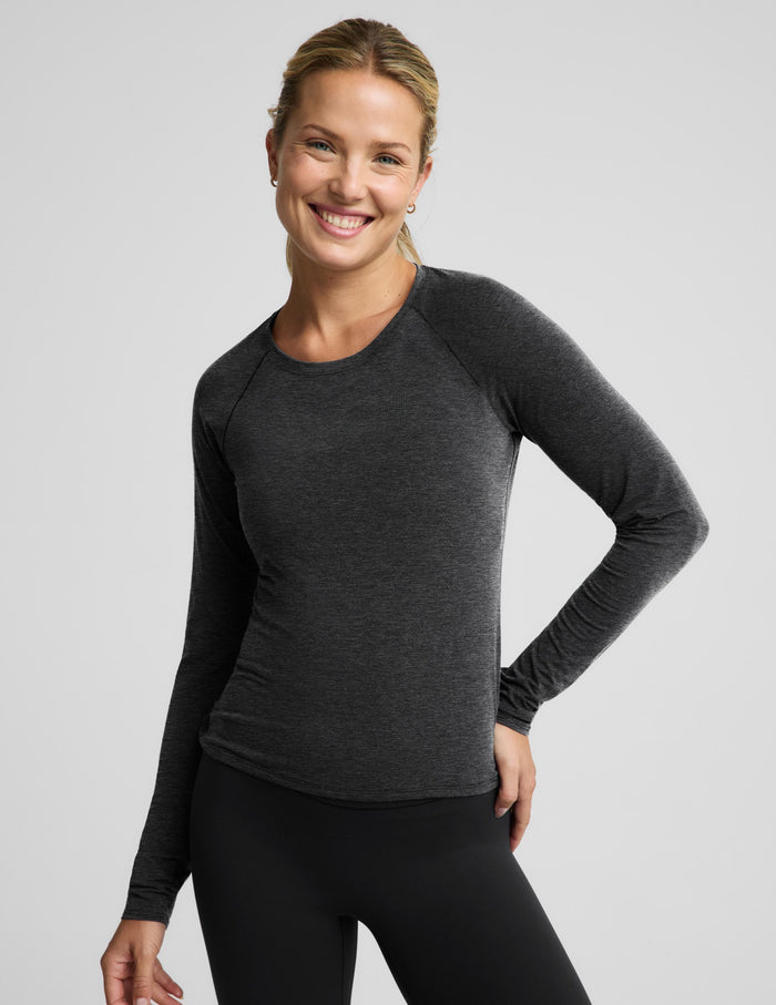 A woman modeling a dark heather gray long-sleeve athletic top with black leggings against a light gray studio background.