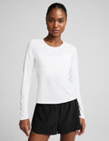 A woman modeling a white long-sleeve crewneck top paired with black athletic shorts, photographed against a solid light gray studio background.