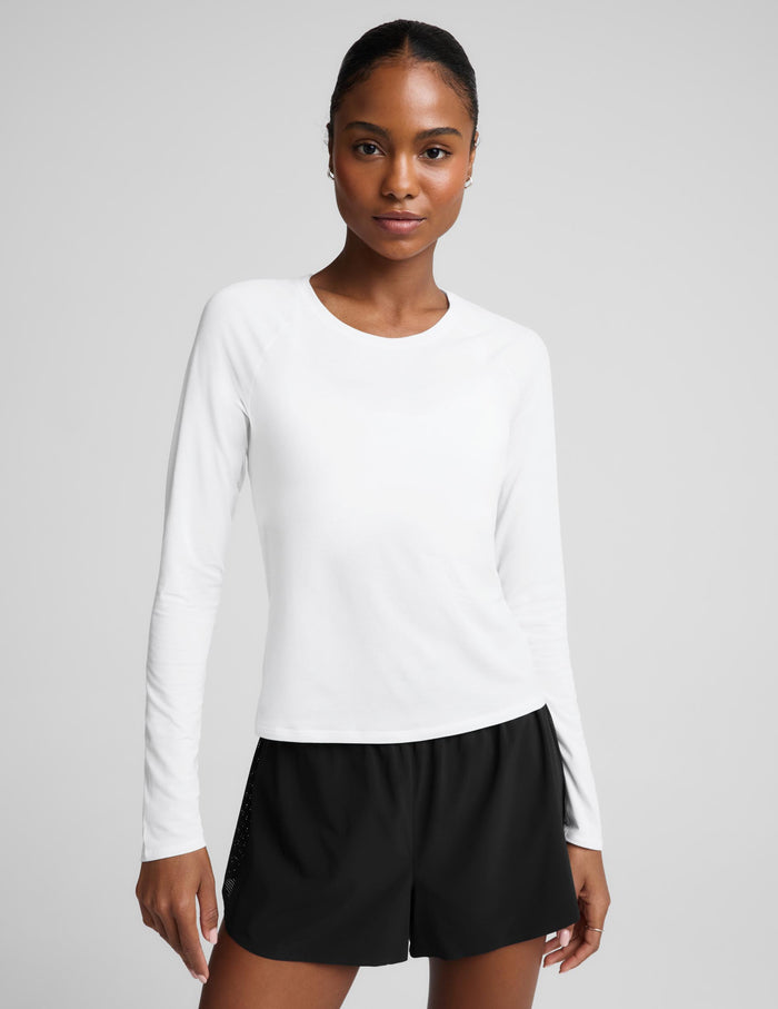 A woman modeling a white long-sleeve crewneck top paired with black athletic shorts, photographed against a solid light gray studio background.