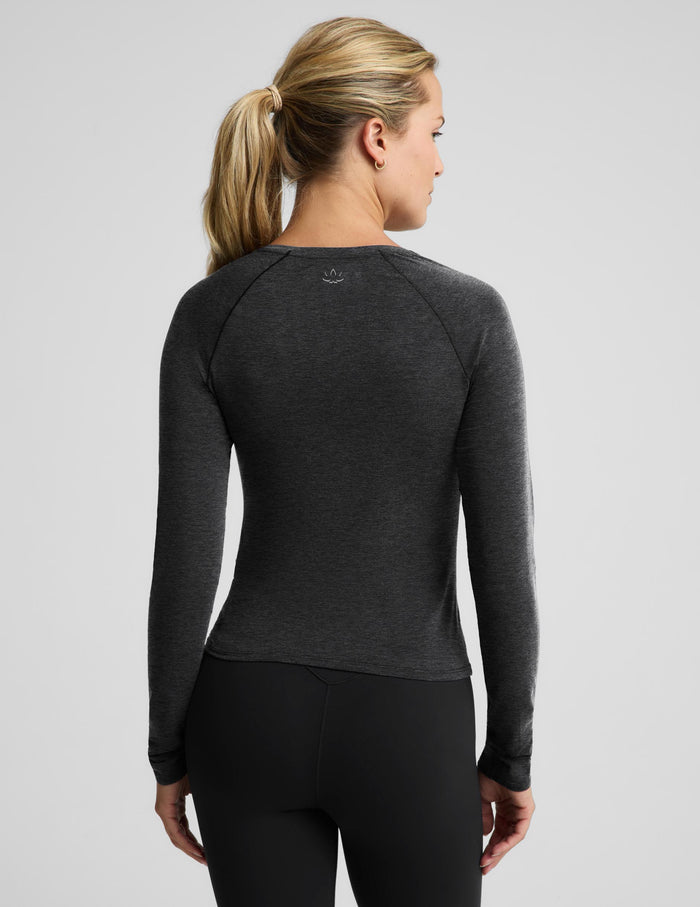 A woman with blonde hair in a ponytail is shown from the back wearing a dark heather gray long-sleeve athletic top and black leggings against a light gray studio background.