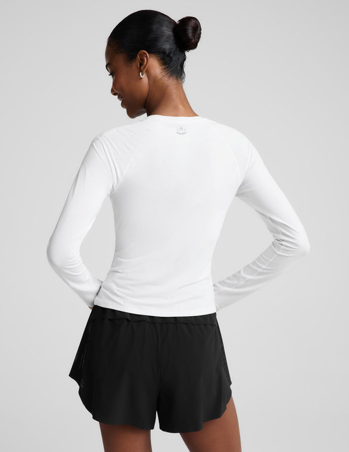 A woman modeling a white long-sleeve crewneck top paired with black athletic shorts, photographed against a solid light gray studio background.