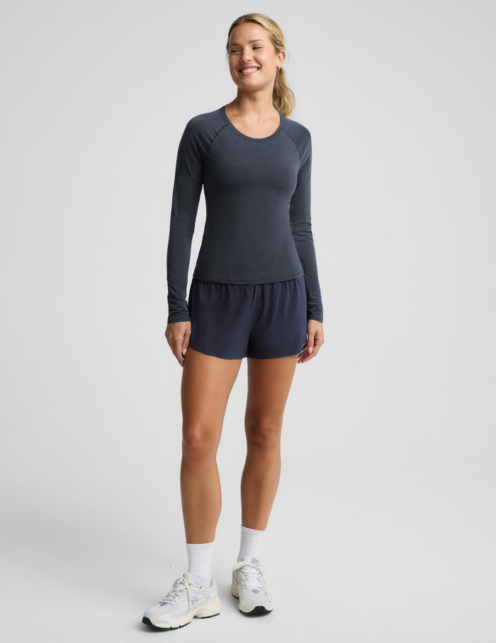 A woman modeling a dark slate blue long-sleeve athletic top and matching shorts, paired with white socks and sneakers, against a light gray studio background.