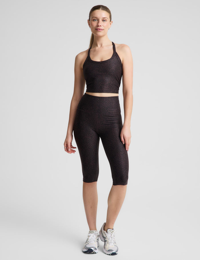 A woman modeling a matching activewear set consisting of a dark brown leopard crop tank and high-waisted pedal pushers, paired with white athletic sneakers, against a light gray studio background.