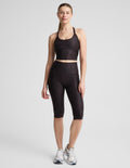 A woman modeling a matching activewear set consisting of a dark brown leopard crop tank and high-waisted pedal pushers, paired with white athletic sneakers, against a light gray studio background.