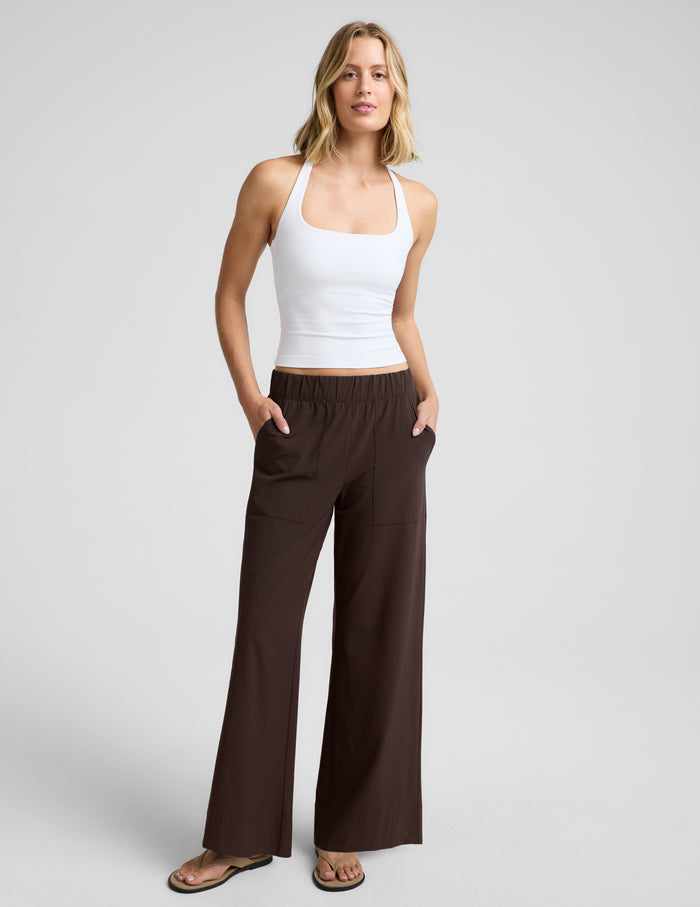 Woman wearing a white halter tank top and brown wide leg pants against a plain studio background.