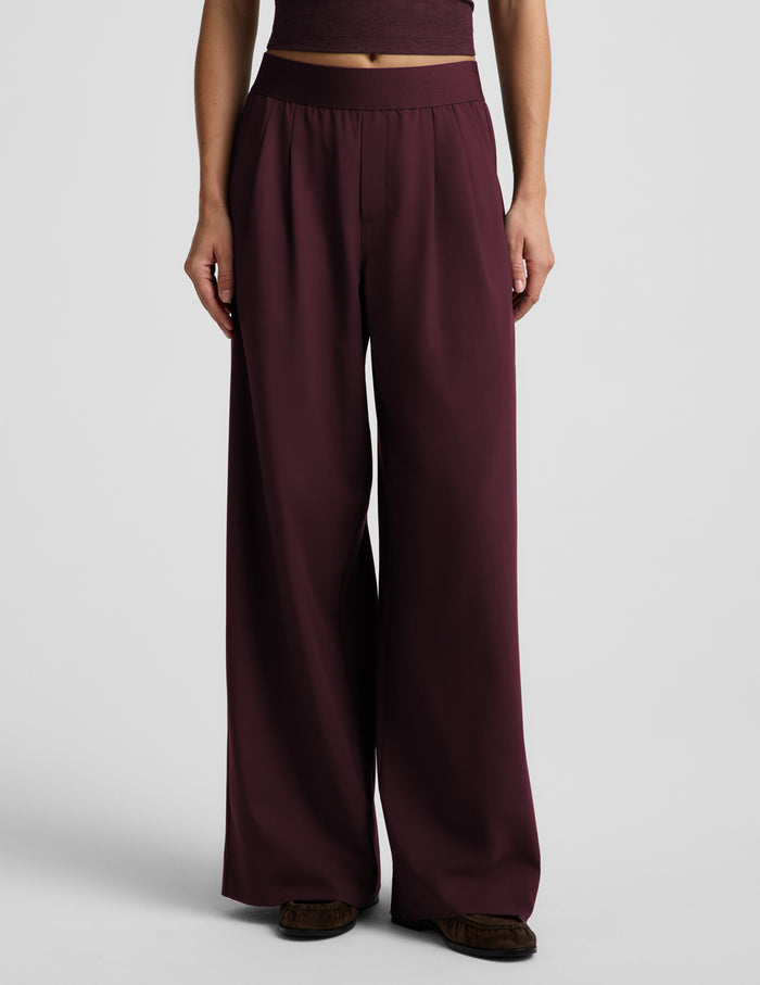 A model wearing a pair of wide-leg, high-waisted trousers in a deep burgundy or plum color, paired with a matching tank top and dark brown slippers, against a light gray studio background.