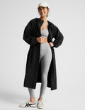 Woman wearing a black trench coat over a gray sports bra and leggings on a plain background.