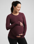 A pregnant woman wearing a long-sleeve crewneck maternity top in maroon, holding her baby bump against a solid light gray background.