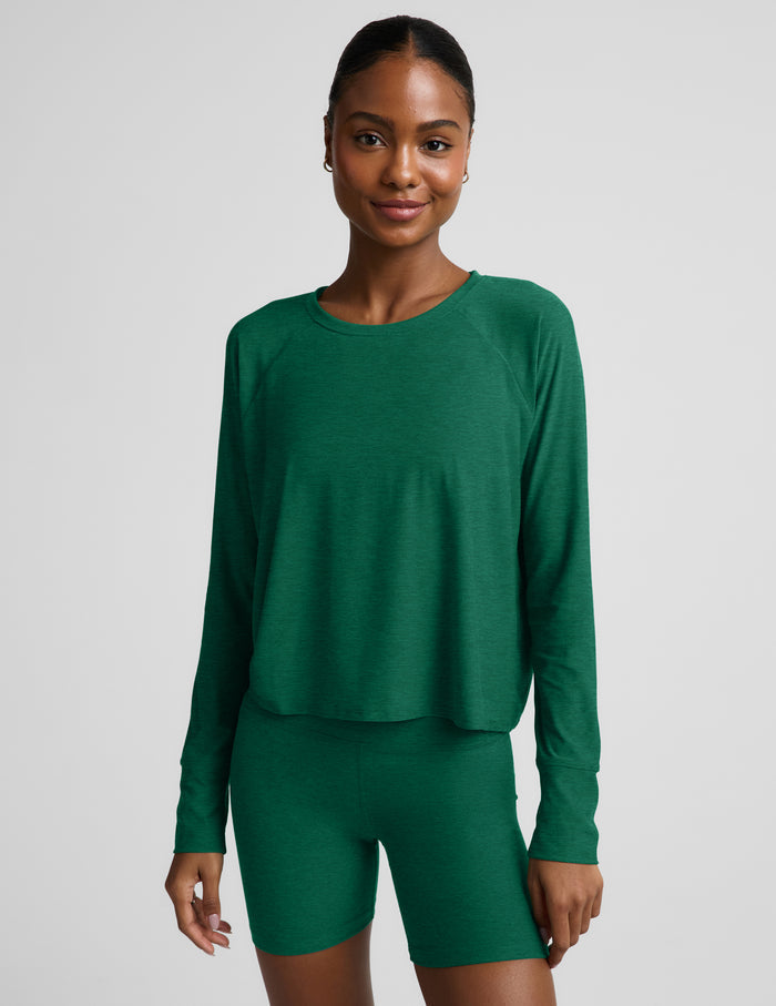 Woman wearing a green long sleeve top and biker shorts against a plain background.