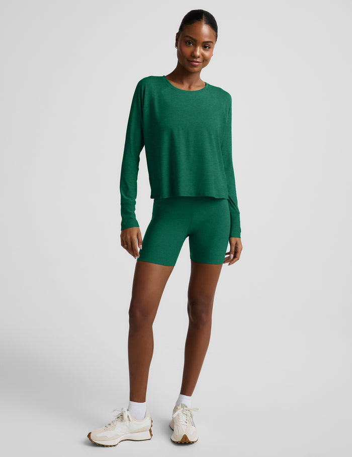 Woman wearing a green long sleeve top and biker shorts against a plain background.