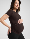 Pregnant woman wearing a brown maternity t-shirt and black leggings against a plain background.