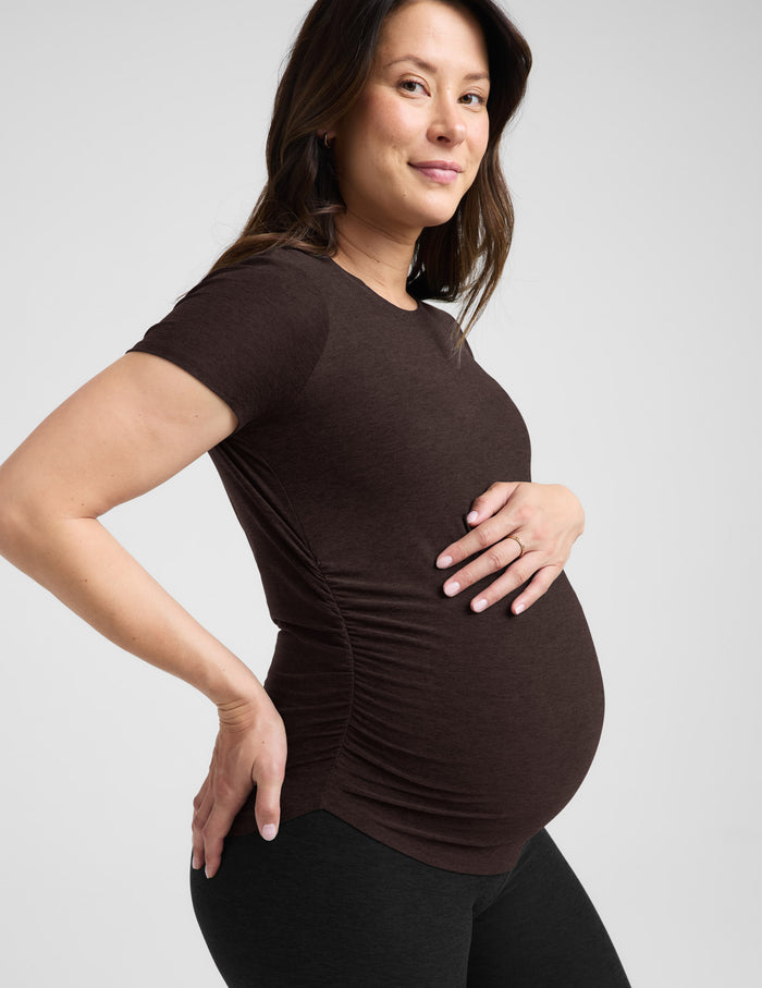 Pregnant woman wearing a brown maternity t-shirt and black leggings against a plain background.