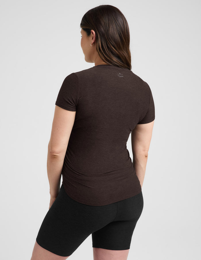 Pregnant woman wearing a brown maternity t-shirt and black leggings against a plain background.