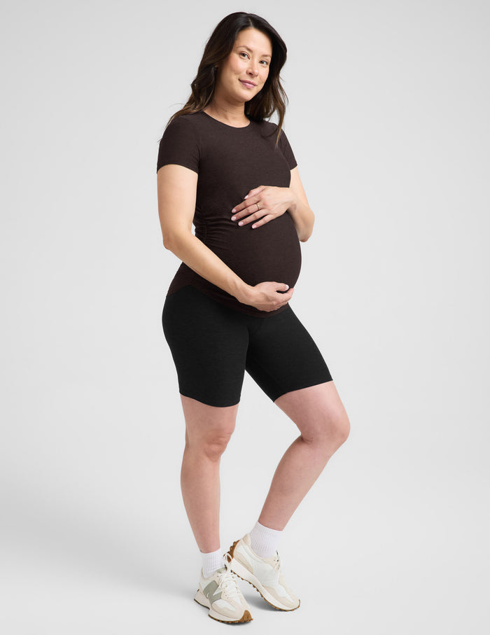 Pregnant woman wearing a brown maternity t-shirt and black leggings against a plain background.