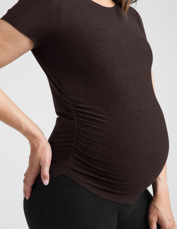 Pregnant woman wearing a brown maternity t-shirt and black leggings against a plain background.