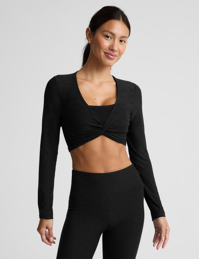 A woman modeling a black long-sleeve crop top with a twisted front detail and matching black leggings against a light gray studio background.