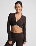 A woman wearing a cropped long sleeve top and brown foldover bootcut pants against a plain background.