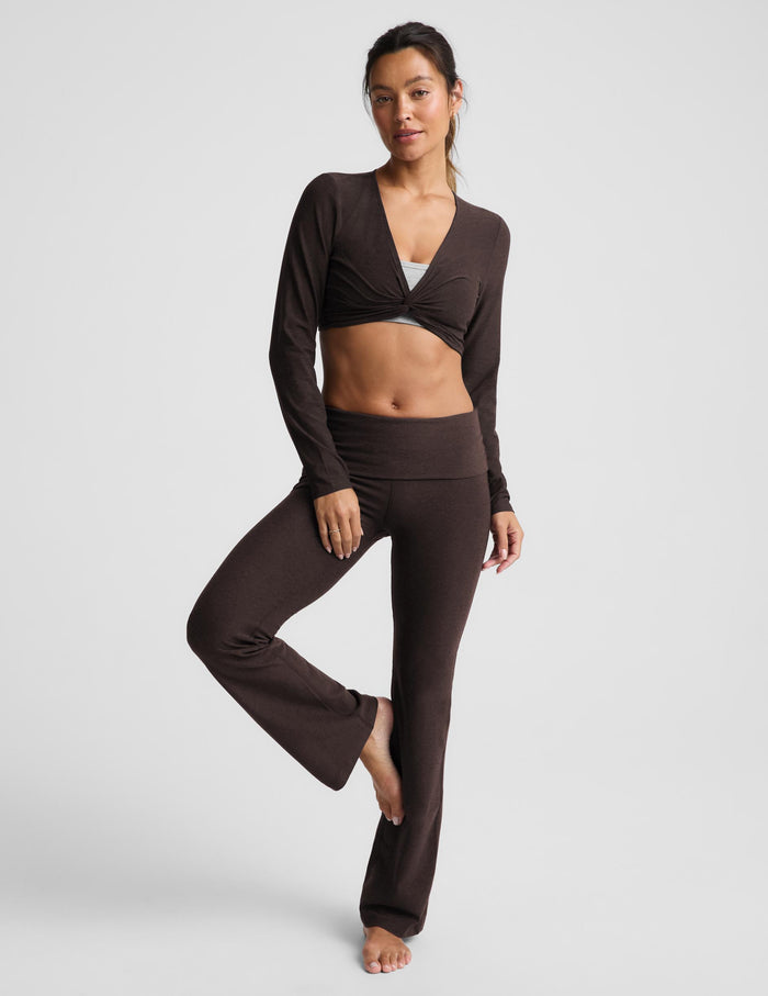 A woman wearing a cropped long sleeve top and brown foldover bootcut pants against a plain background.