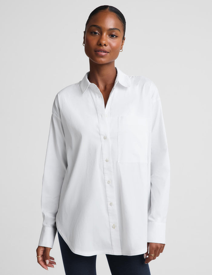 A woman modeling a white long-sleeve button-up shirt with a chest pocket, paired with dark pants, against a light gray studio background.
