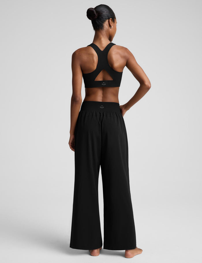 A woman modeling a black two-piece activewear set consisting of a bra and high-waisted wide-leg pants, photographed against a solid light gray studio background.