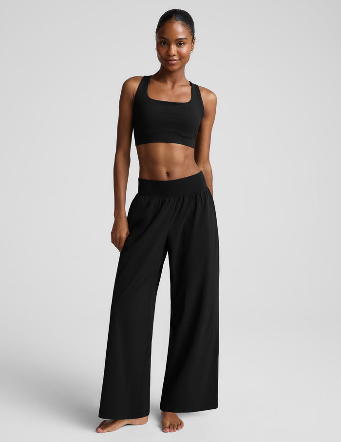 A woman modeling a black two-piece activewear set consisting of a cropped tank top and high-waisted wide-leg pants, photographed against a solid light gray studio background.