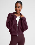 A woman wearing a cropped, zip-up fleece jacket in a deep burgundy color, styled over a white t-shirt and matching burgundy leggings, against a light gray studio background.