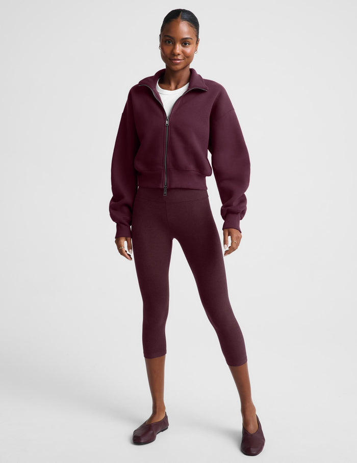 A woman wearing a cropped, zip-up fleece jacket in a deep burgundy color, styled over a white t-shirt and matching burgundy leggings, against a light gray studio background.