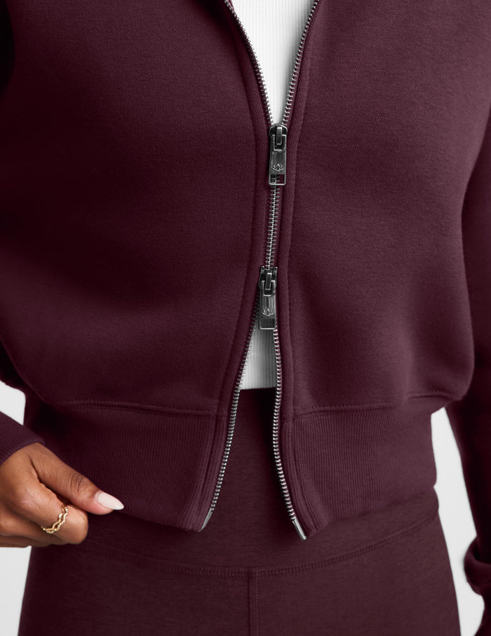 A woman wearing a cropped, zip-up fleece jacket in a deep burgundy color, styled over a white t-shirt and matching burgundy leggings, against a light gray studio background.