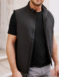 Man wearing a black vest over a black shirt with a neutral background.
