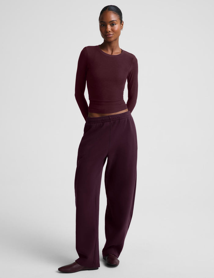 A model wearing a matching dark burgundy two-piece lounge set consisting of a long-sleeve top and wide-leg sweatpants, photographed against a neutral light gray studio background.