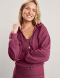 A smiling blonde woman wearing a mauve-red zip-up hoodie and matching leggings against a light gray studio background.