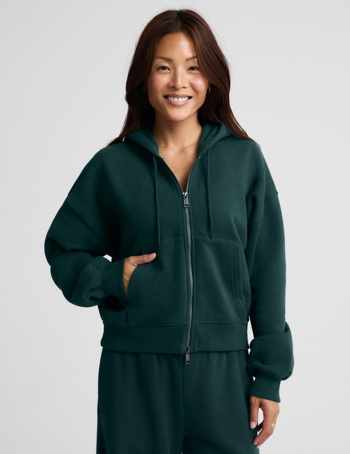 A woman wearing a dark forest green full-zip hoodie and matching sweatpants, standing against a light gray studio background.