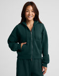 A woman wearing a dark forest green full-zip hoodie and matching sweatpants, standing against a light gray studio background.