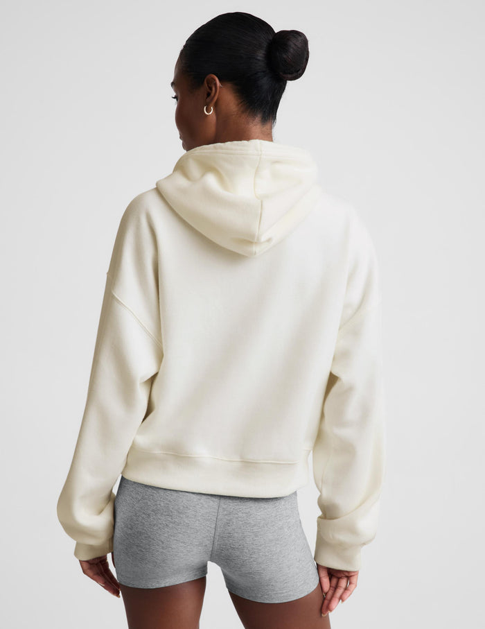 A woman modeling a cream-colored zip-up hoodie with a relaxed fit, paired with a gray sports bra and heather gray bike shorts, against a solid light gray background.