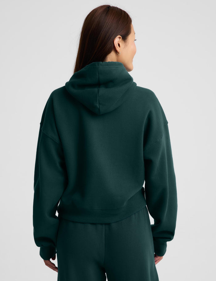 A woman wearing a dark forest green full-zip hoodie and matching sweatpants, standing against a light gray studio background.
