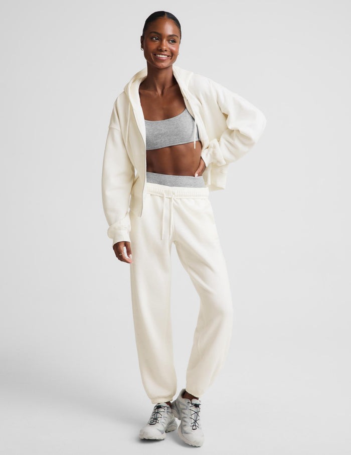 A woman modeling a matching cream-colored lounge set consisting of a zip-up hoodie and jogger pants, styled with a gray sports bra and white sneakers against a light gray studio background.