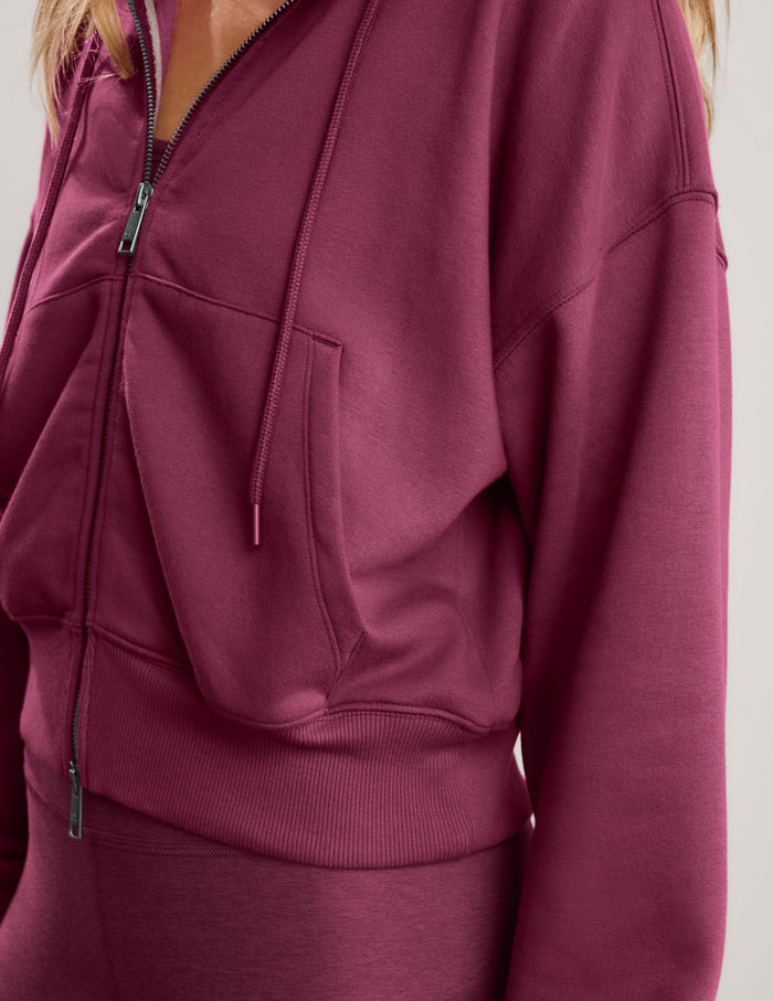 A blonde woman wearing a mauve-red zip-up hoodie and matching leggings against a light gray studio background.