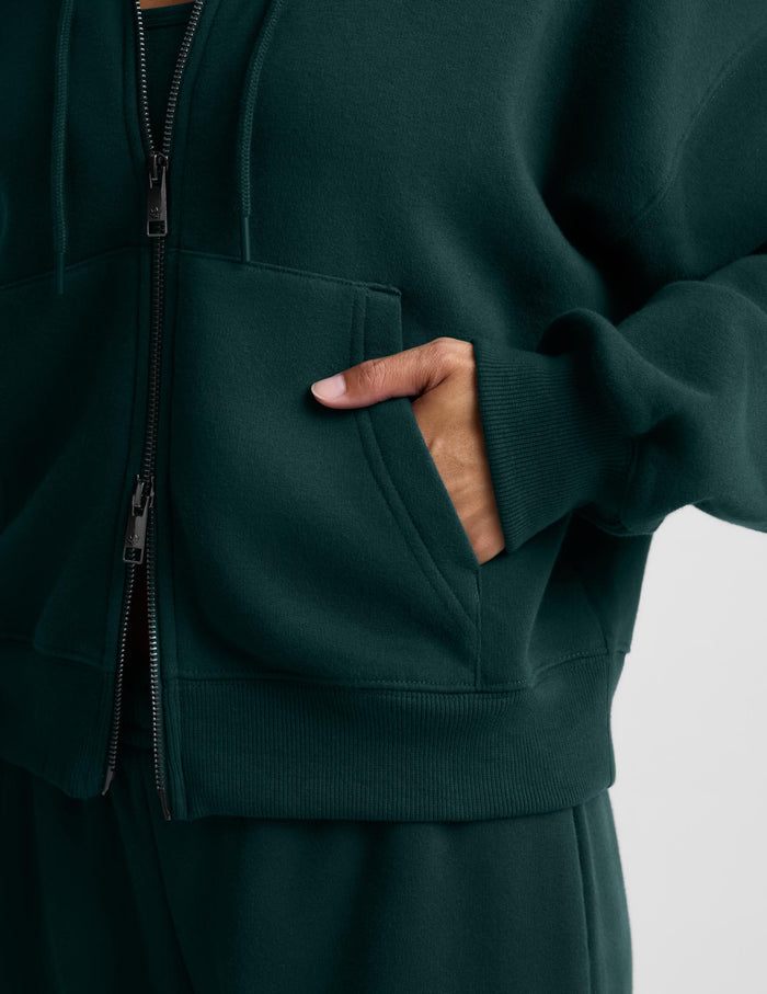 A woman wearing a dark forest green full-zip hoodie and matching sweatpants, standing against a light gray studio background.