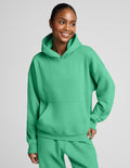 A woman wearing a bright green pullover hoodie and matching sweatpants, photographed against a plain light gray studio background.
